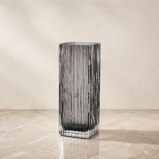 A tall, rectangular ribbed glass vase in a smoky translucent gray color, standing on a beige marble surface against a textured beige wall.