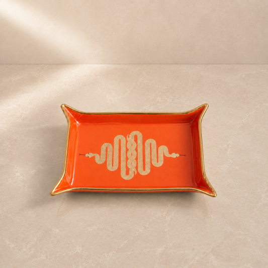 An orange rectangular ceramic tray with notched corners, a gold rim, and a cream-colored snake motif in the center, set against a beige marble-textured background.