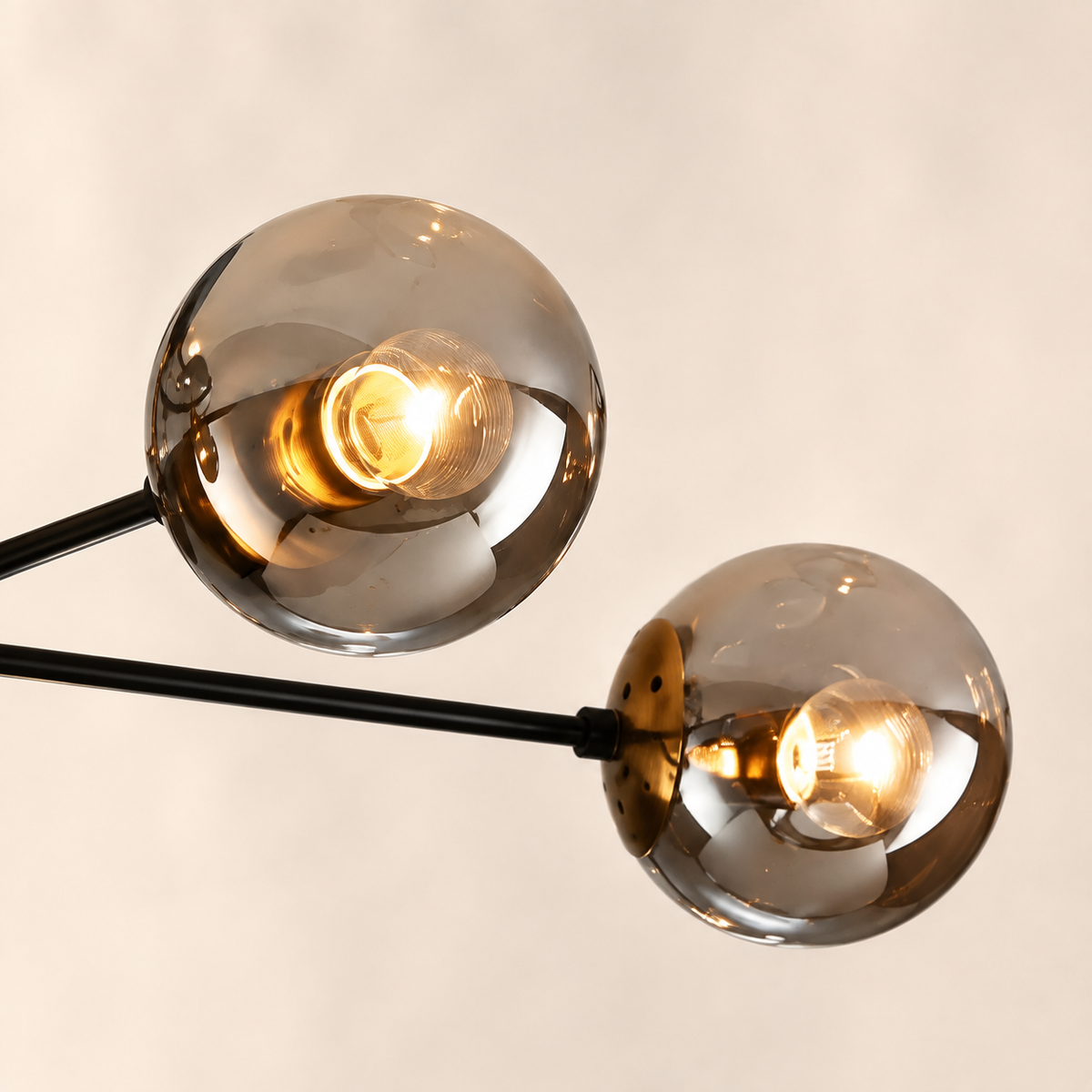 A close-up of a modern two-light sputnik chandelier with spherical smoked glass shades and visible warm Edison bulbs on black metal arms, set against a plain light beige background.