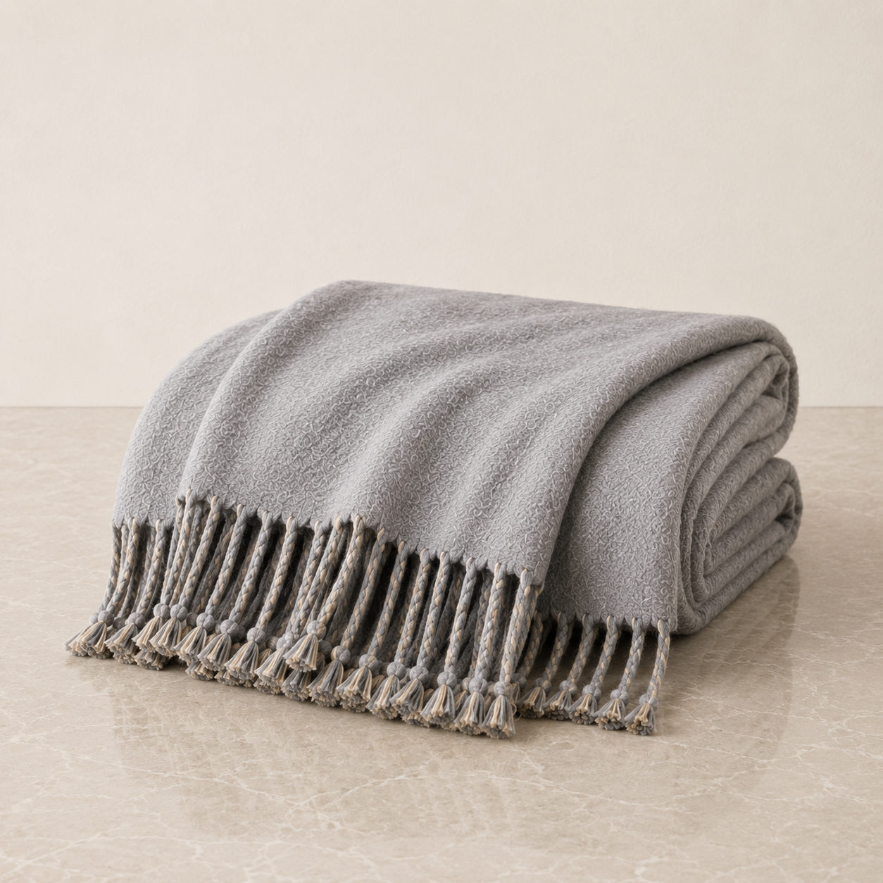 A folded light gray knit throw blanket with braided fringe tassels, displayed on a light beige marble surface against a neutral beige background.