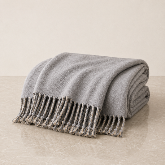 A folded light gray knit throw blanket with braided fringe tassels, displayed on a light beige marble surface against a neutral beige background.