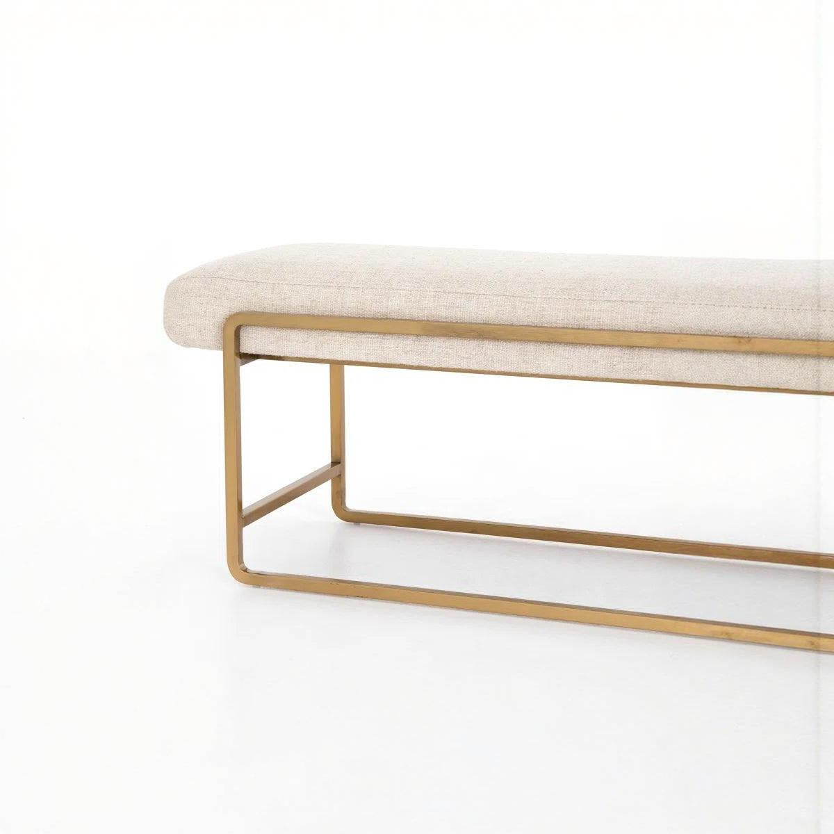 Beige upholstered bench with rounded edges and brass-finished steel frame on a white background