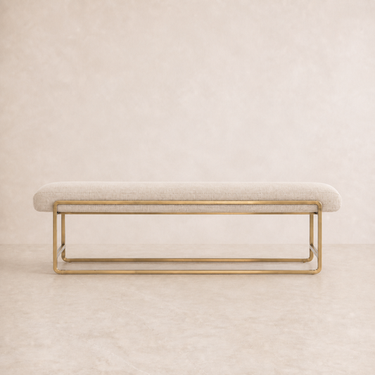 A modern upholstered bench with a light beige fabric seat and a polished gold metal frame, set against a matching beige textured background.