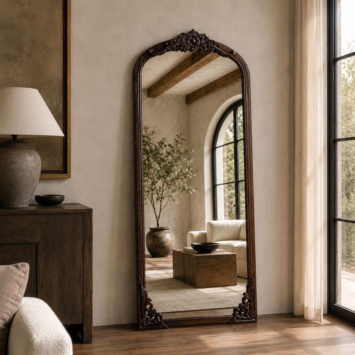 wall mirror with ornate dark carved wood trim stands in a styled room with beige walls, a wooden cabinet, a lamp, and a window looking out onto greenery.
