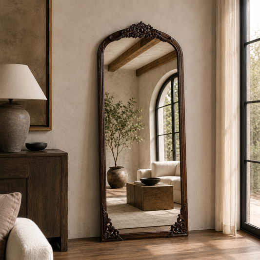 wall mirror with ornate dark carved wood trim stands in a styled room with beige walls, a wooden cabinet, a lamp, and a window looking out onto greenery.