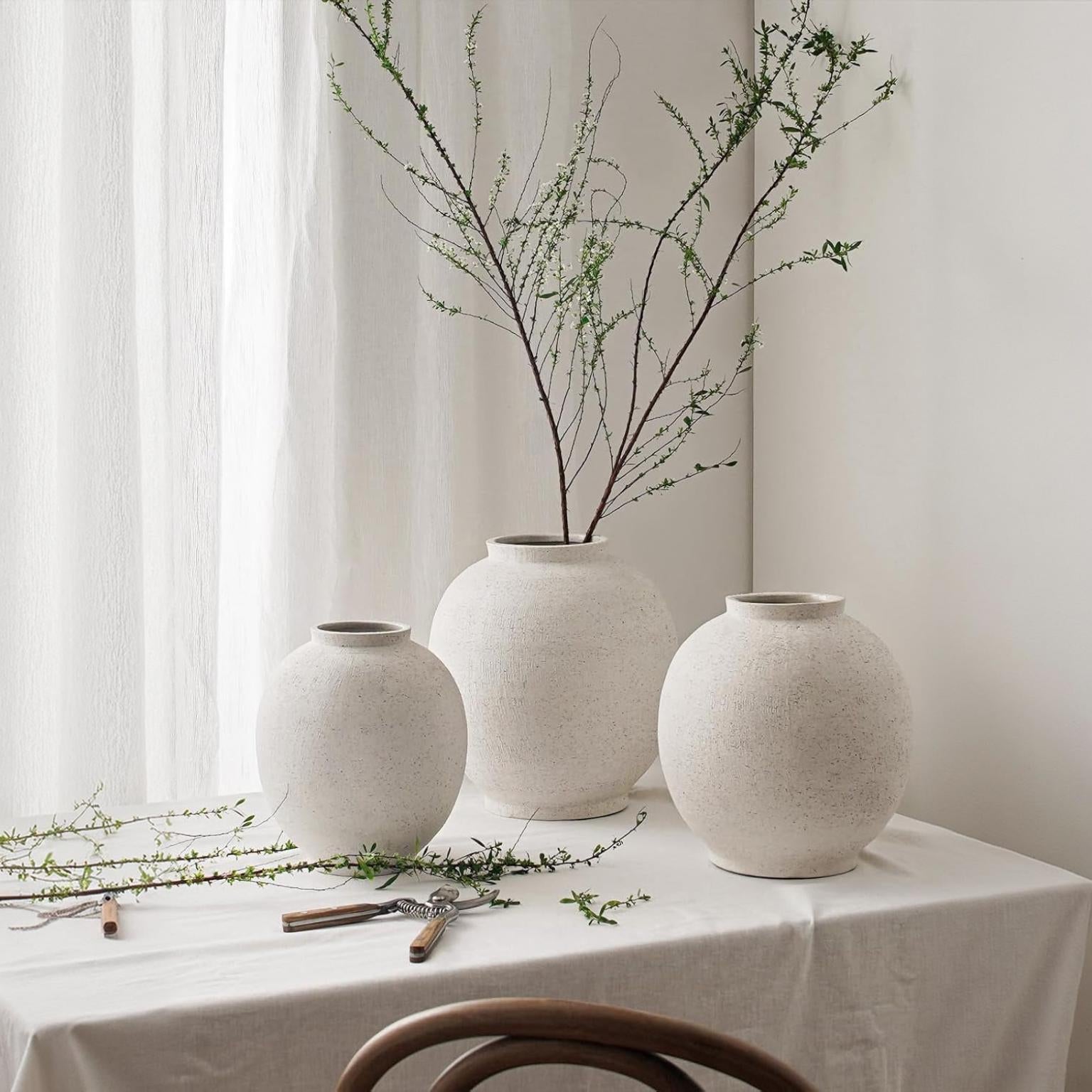 Three large white ceramic vases with rounded shapes displayed on a linen-covered table by a window