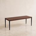 Load image into Gallery viewer, A rectangular wicker bench with a brown woven leather seat and black metal legs, photographed in a minimalist studio setting with a beige wall and floor.

