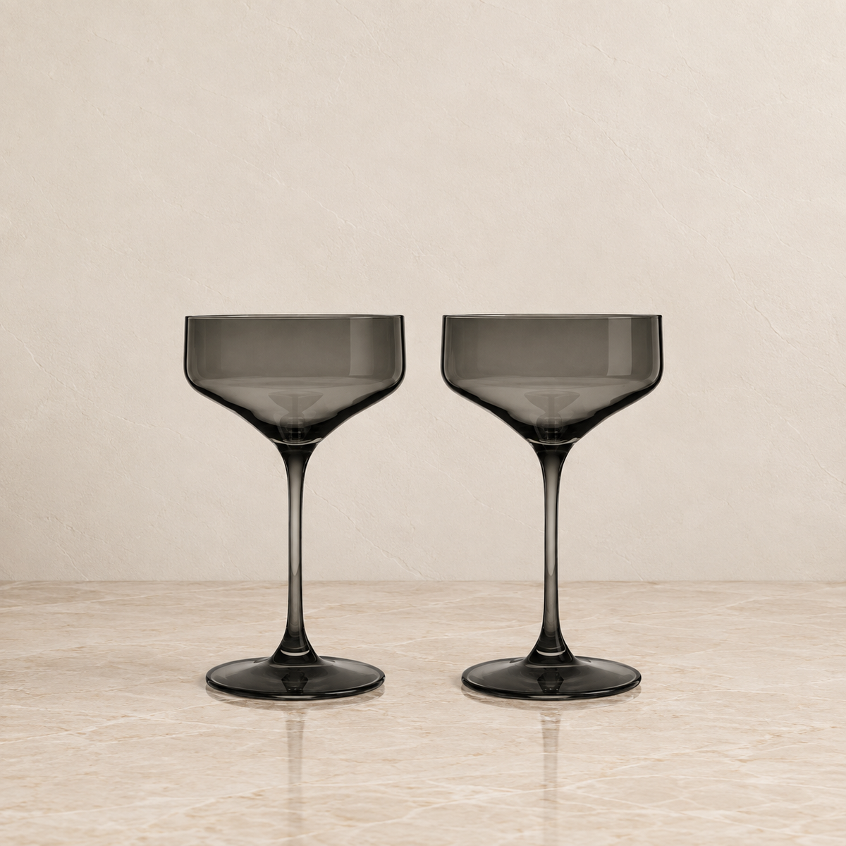 Two dark gray smoked glass coupe glasses on a light beige marble surface against a textured beige background.