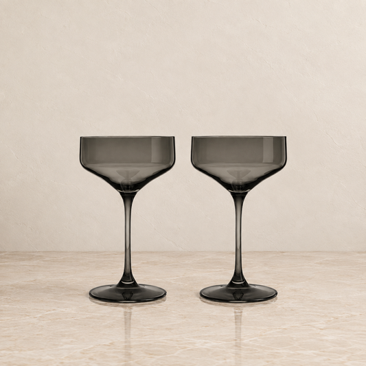Two dark gray smoked glass coupe glasses on a light beige marble surface against a textured beige background.