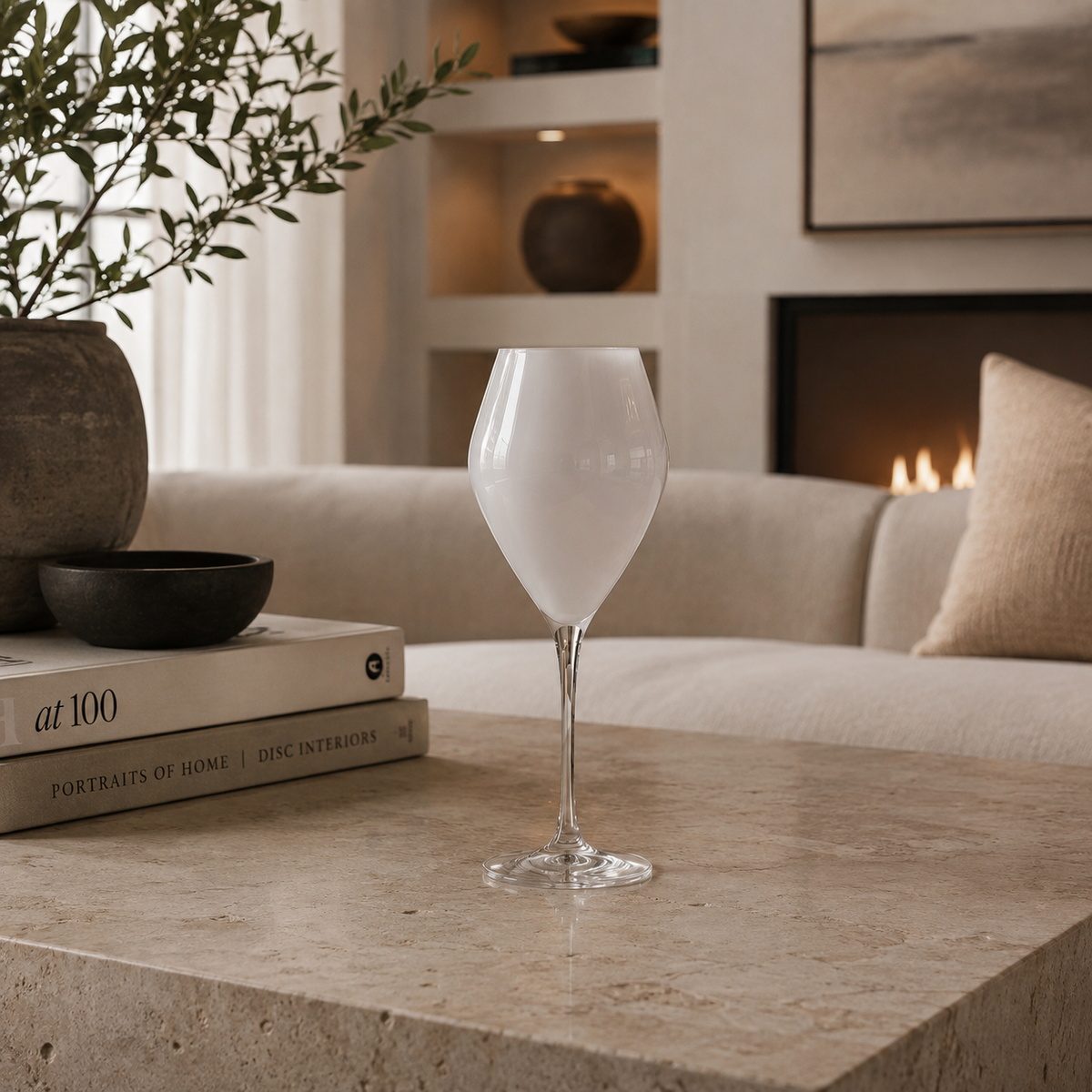 A clear wine glass with a frosted white body sits on a beige stone table in a modern living room setting with a sofa, fireplace, and books in the background.