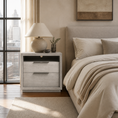 Load image into Gallery viewer, A modern bedroom scene featuring a light gray two-drawer nightstand beside a bed with beige bedding and a textured lamp on top, set in a softly lit room with a window and city view.
