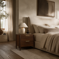 Load image into Gallery viewer, A warm, modern bedroom scene featuring a dark brown wooden nightstand with two drawers and gold-tone handles, styled with a textured ceramic table lamp and a small dark vase. The nightstand is positioned next to a bed with beige linens, pillows, and a textured throw blanket, set against a neutral beige wall with a large abstract painting.
